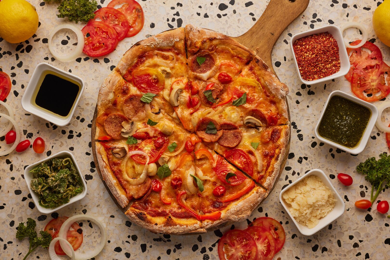 Pizza con distintos ingredientes, rodeada de productos y condimentos sobre una encimera moteada.