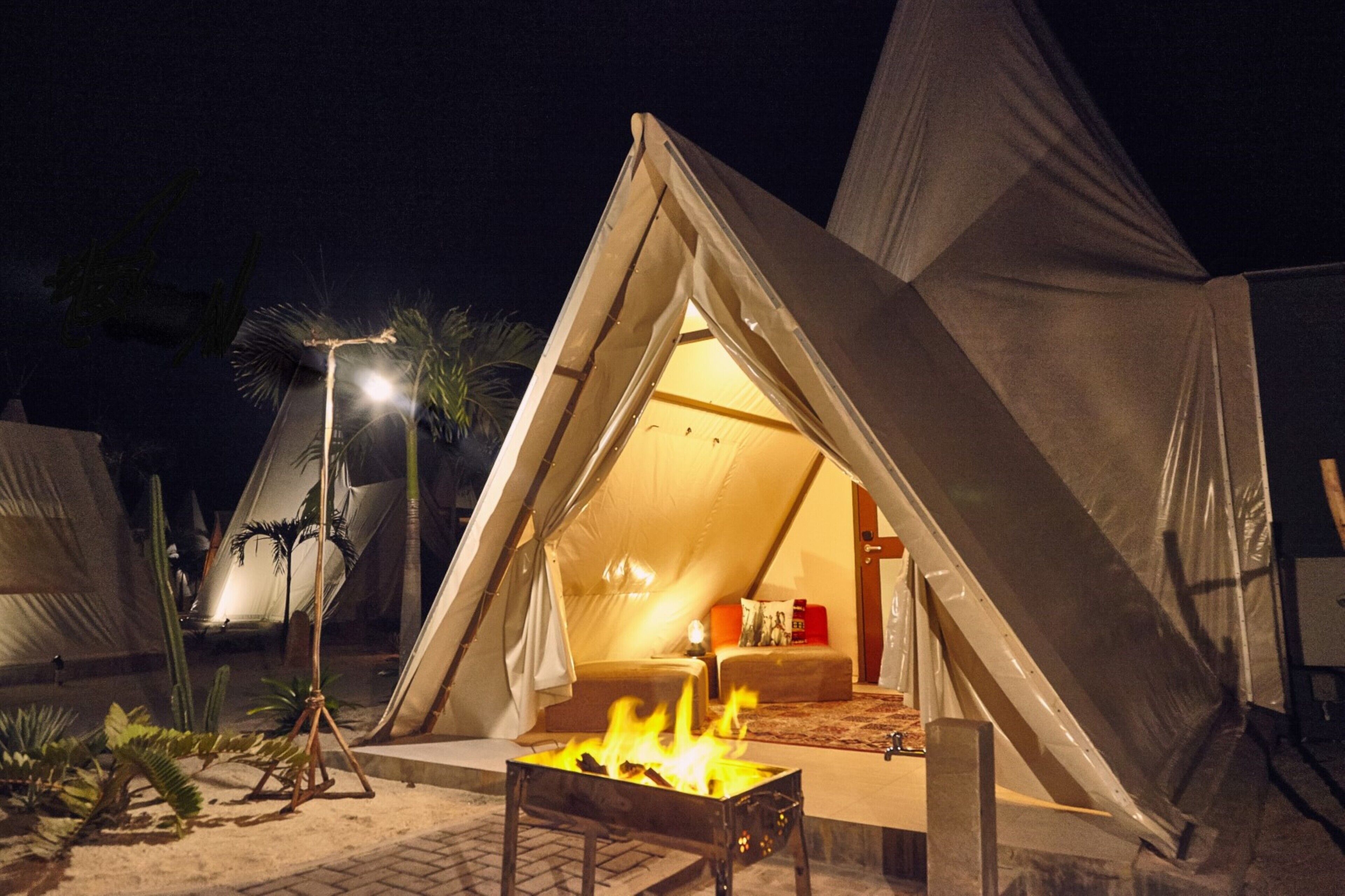 Una carpa con cama en su interior, un hogar de fuego al frente y palmeras alrededor.