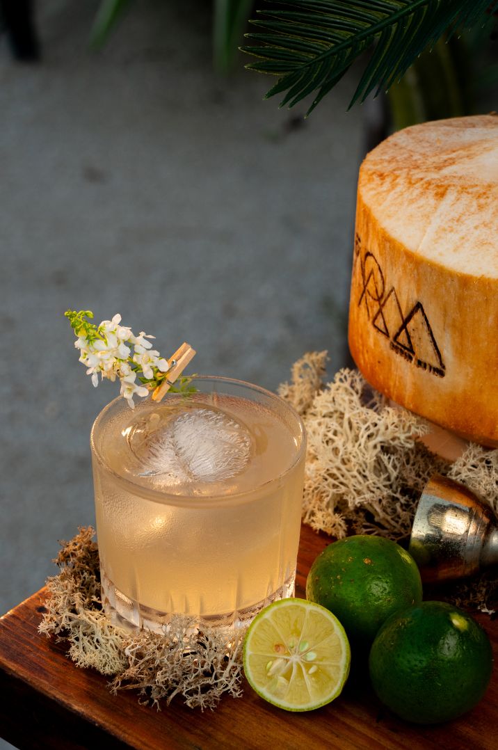 Un vaso de una bebida clara con adorno, mitades de lima y un coco sobre una superficie de madera.