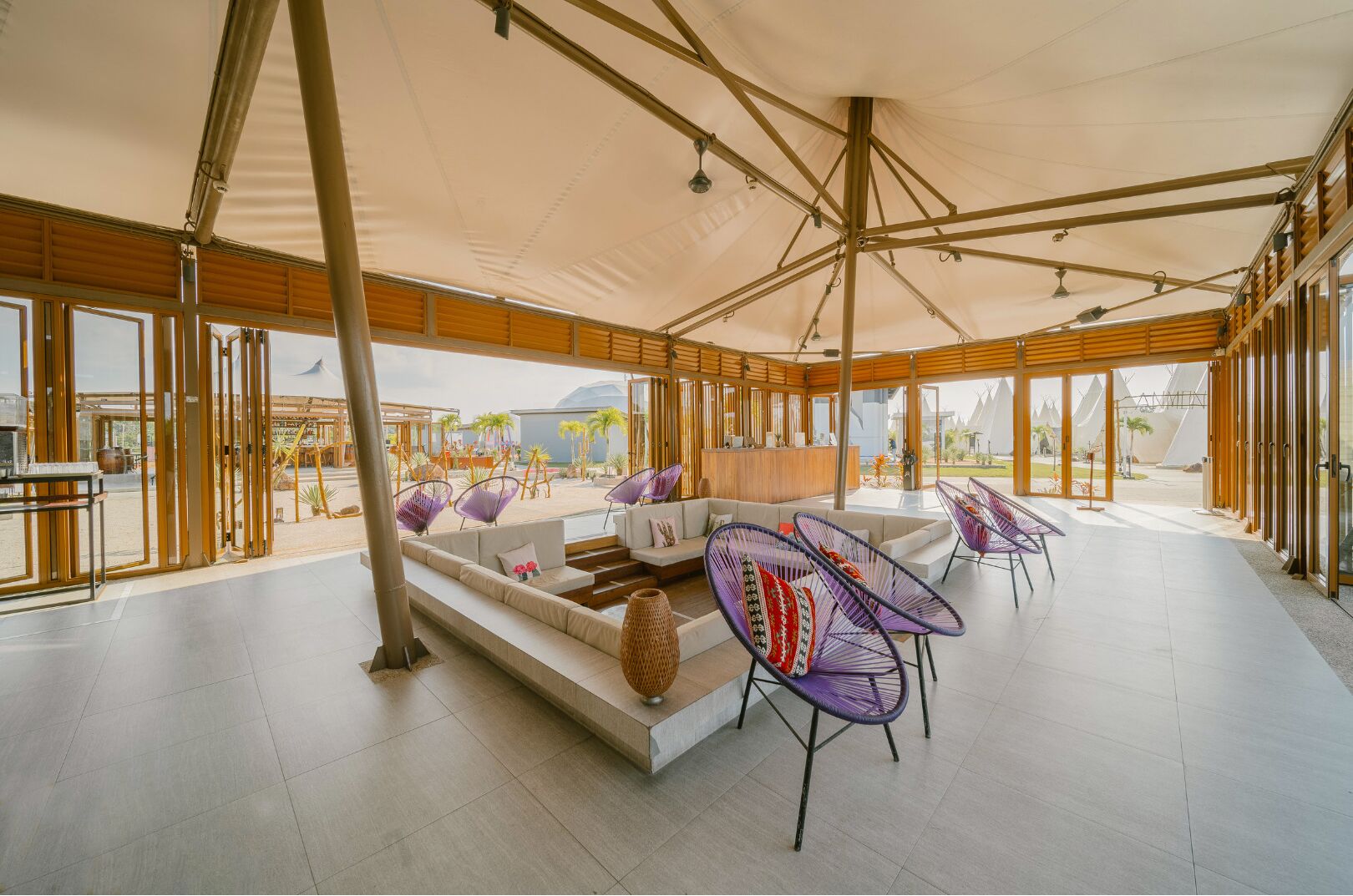 A spacious outdoor lounge with a modern design, featuring a central seating area with sofas and chairs, and large windows offering views of the surrounding landscape.