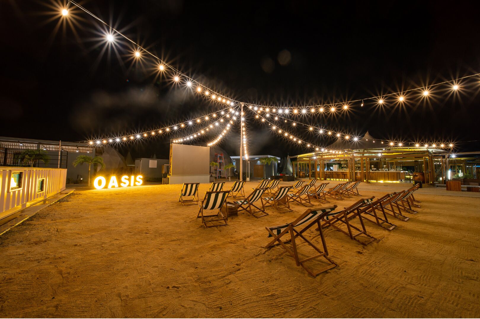 A beachside resort with a bar area, lounge chairs, and string lights.