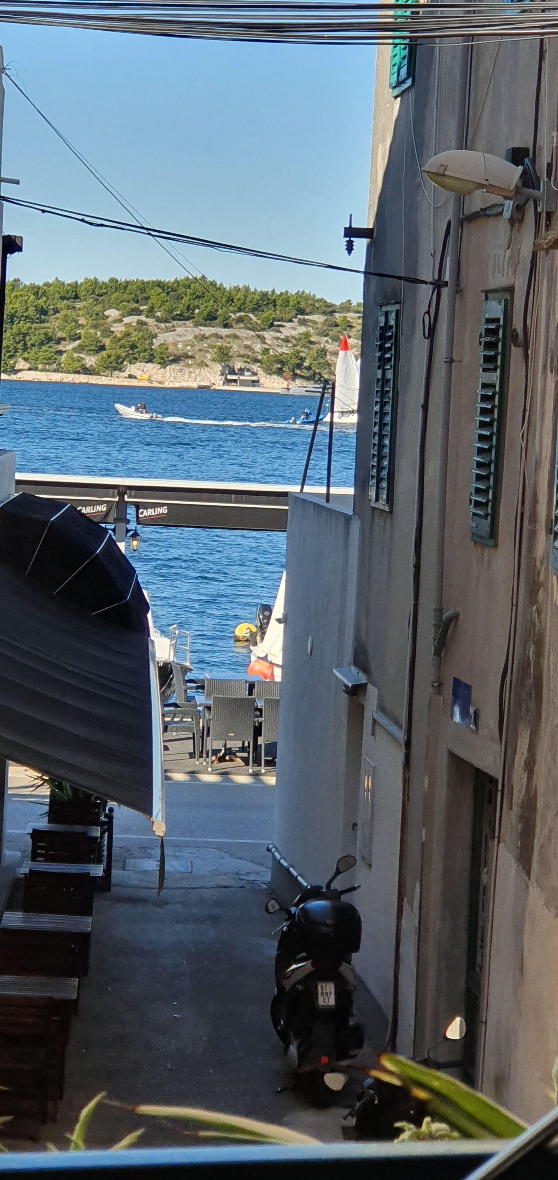 A narrow street with a scooter parked on the side, a boat on the water, and a view of the sea and a distant shoreline.