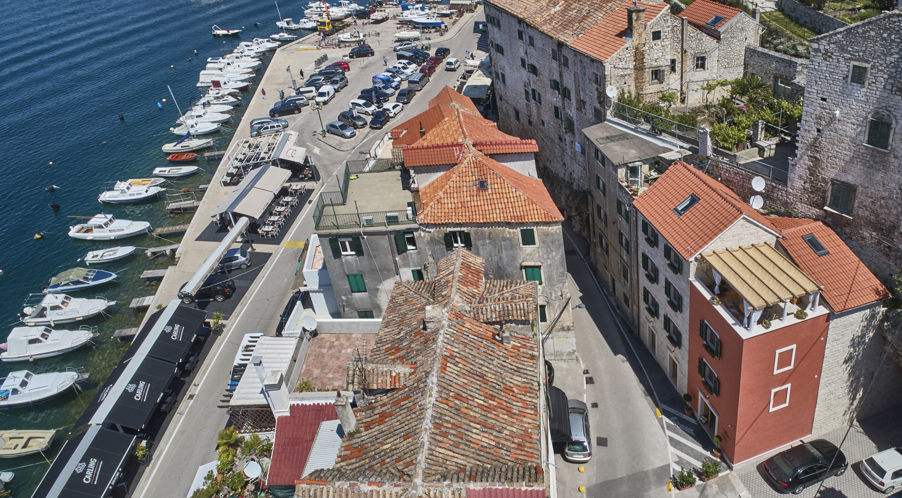 Vista aerea di una cittadina costiera con un porto turistico, edifici in pietra con tetti in tegole rosse e una combinazione di edifici residenziali e commerciali.
