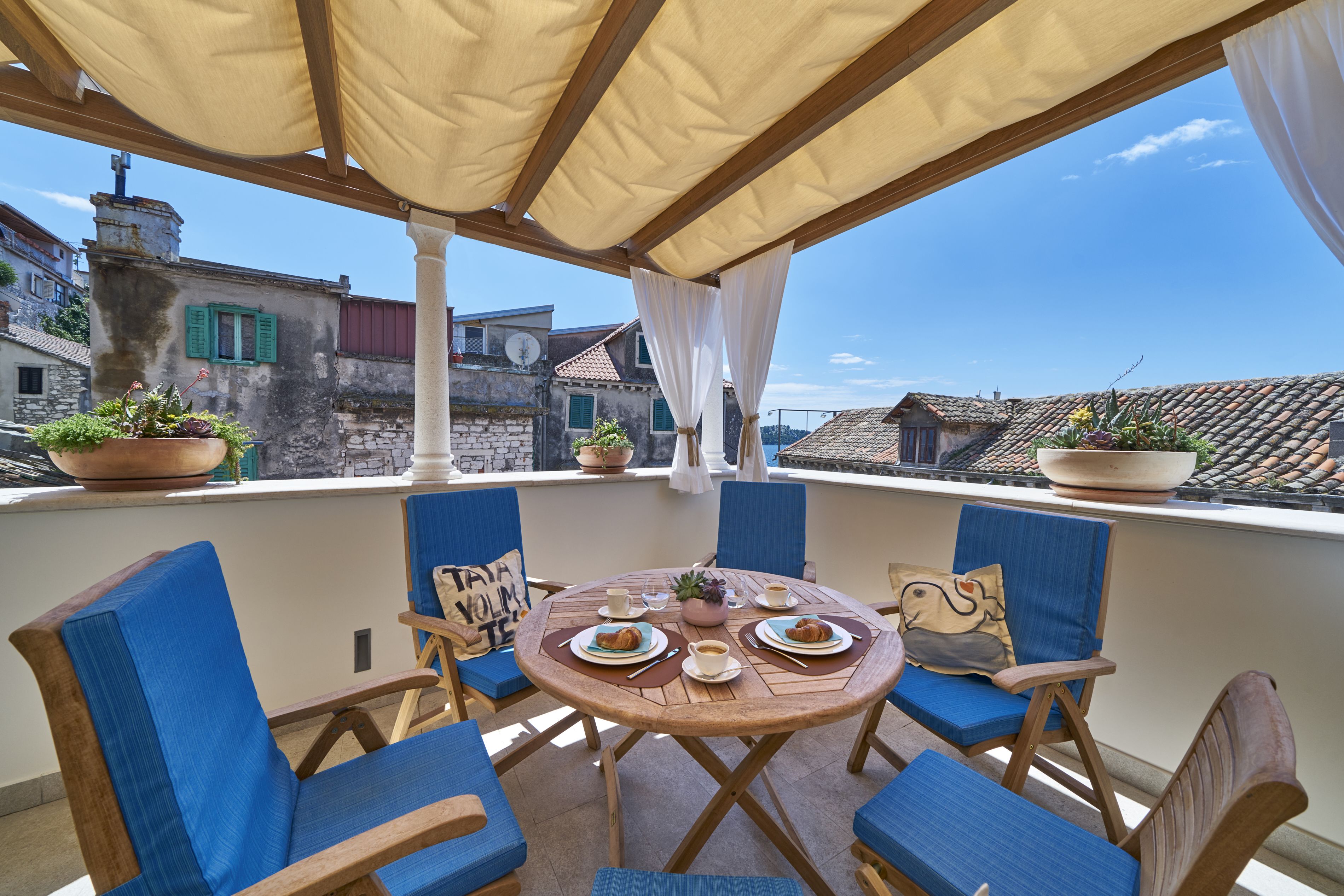 A round wooden table with blue upholstered chairs, a potted plant, and a view of historic buildings.