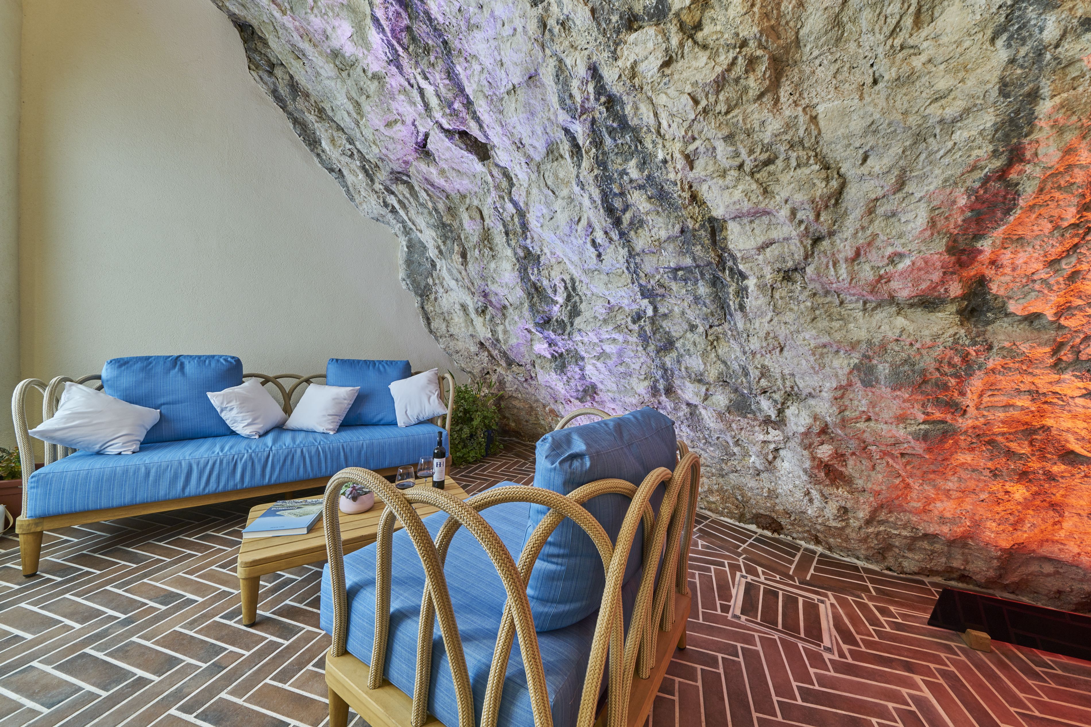A room with a blue sofa, a wicker chair, and a coffee table with a book and bottle.