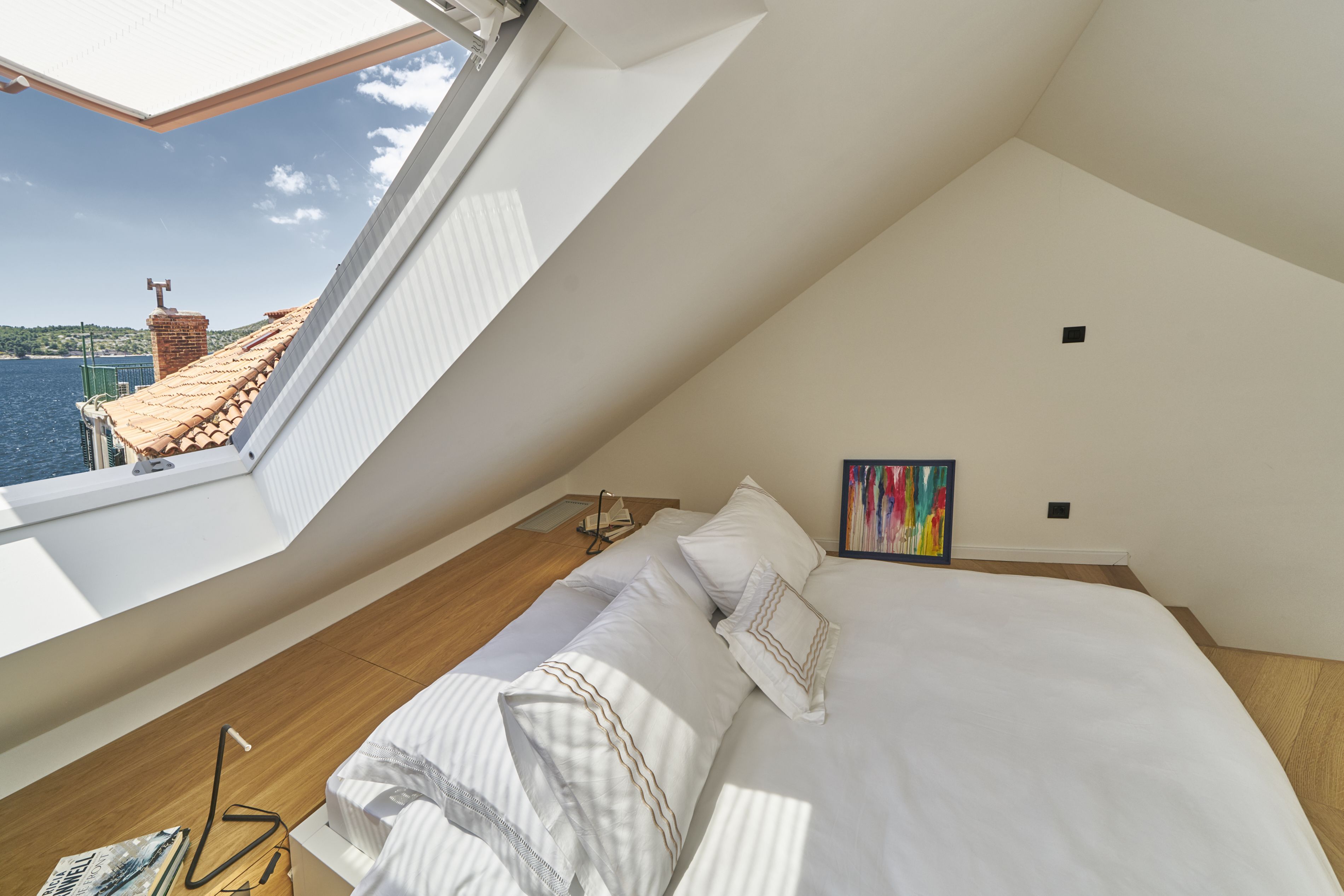 A bedroom with a sloped ceiling, a bed with white linens, a painting on the wall, and a view of the sea through a skylight.
