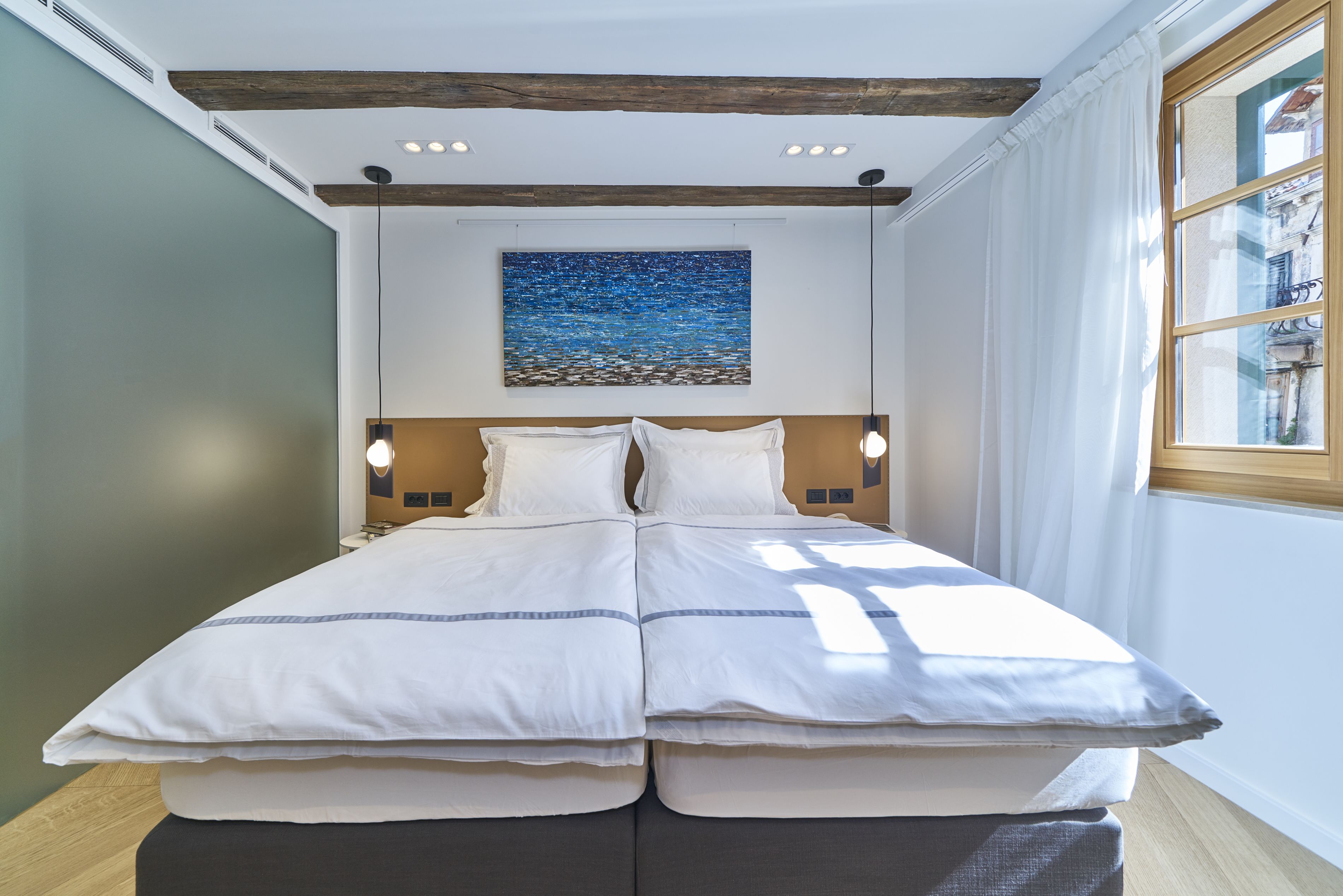 A double bed with white bedding, a wooden headboard, two bedside lamps, and a painting of a blue sky and sea on the wall.