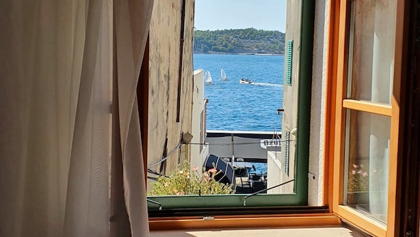 A window with a view of a body of water, a sailboat, and a building with a green door. - Villa Pergola (Sibenik)