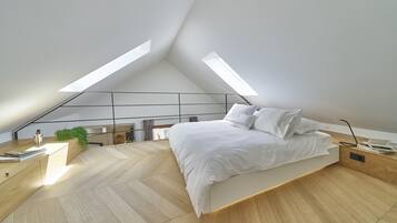 Quarto moderno com teto inclinado, cama com roupa de cama branca, escrivaninha de madeira e uma pequena planta.