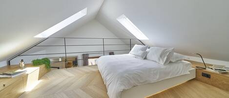 Une chambre moderne avec un plafond en pente, un lit avec une literie blanche, un bureau en bois et une petite plante.