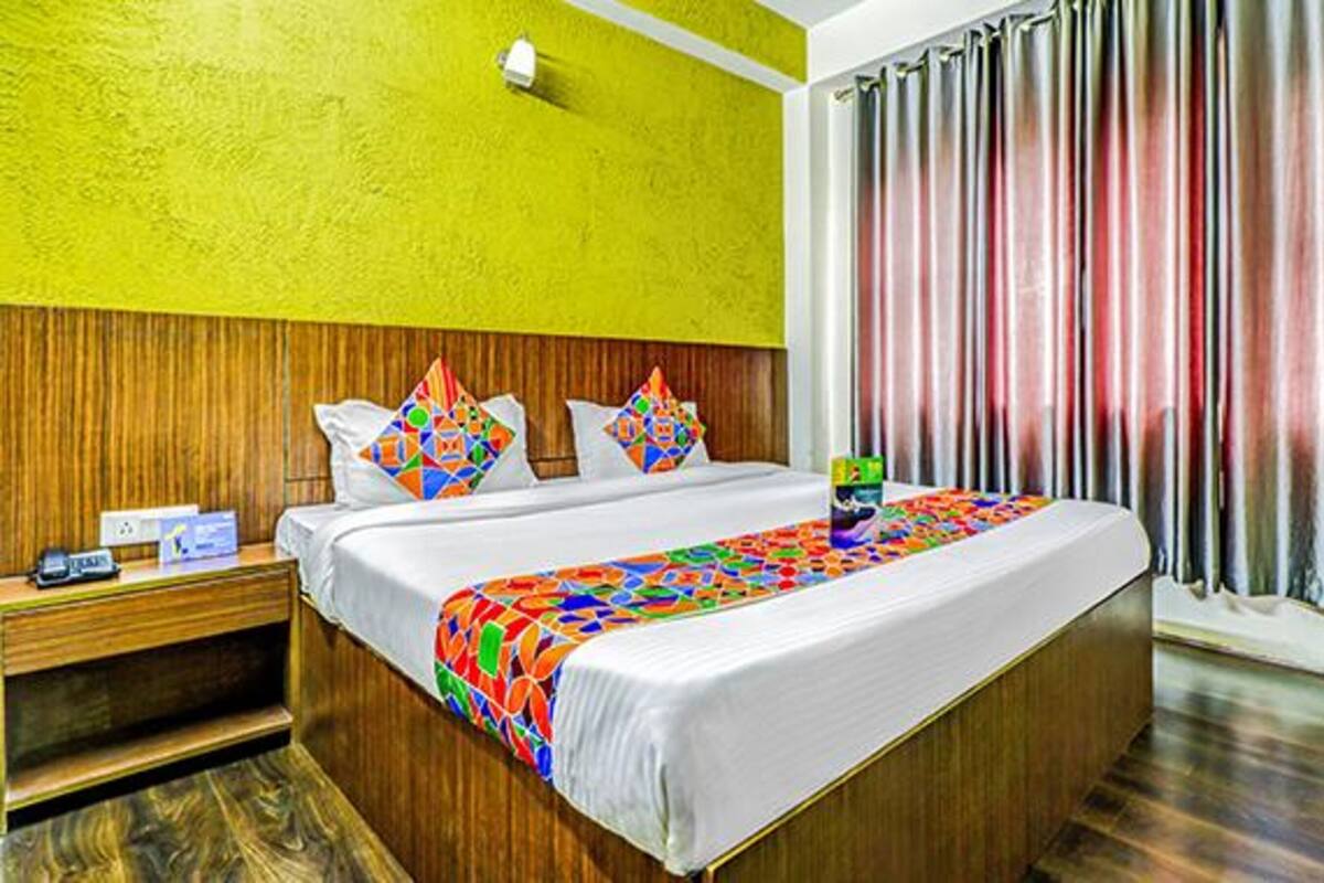 A hotel room with a wooden headboard, a bed with colorful pillows, a bedside table with a phone and a box, and a window with curtains.