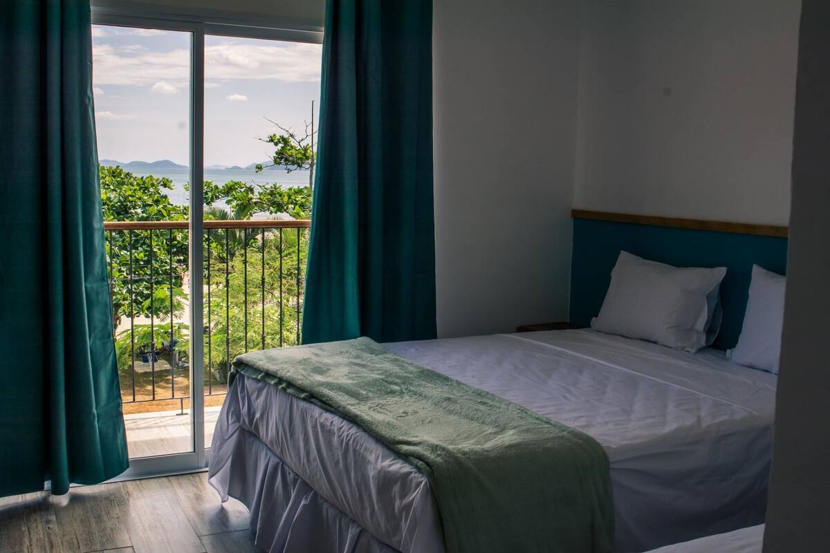 A hotel room with a bed, a balcony with curtains, and a view of greenery and mountains.
