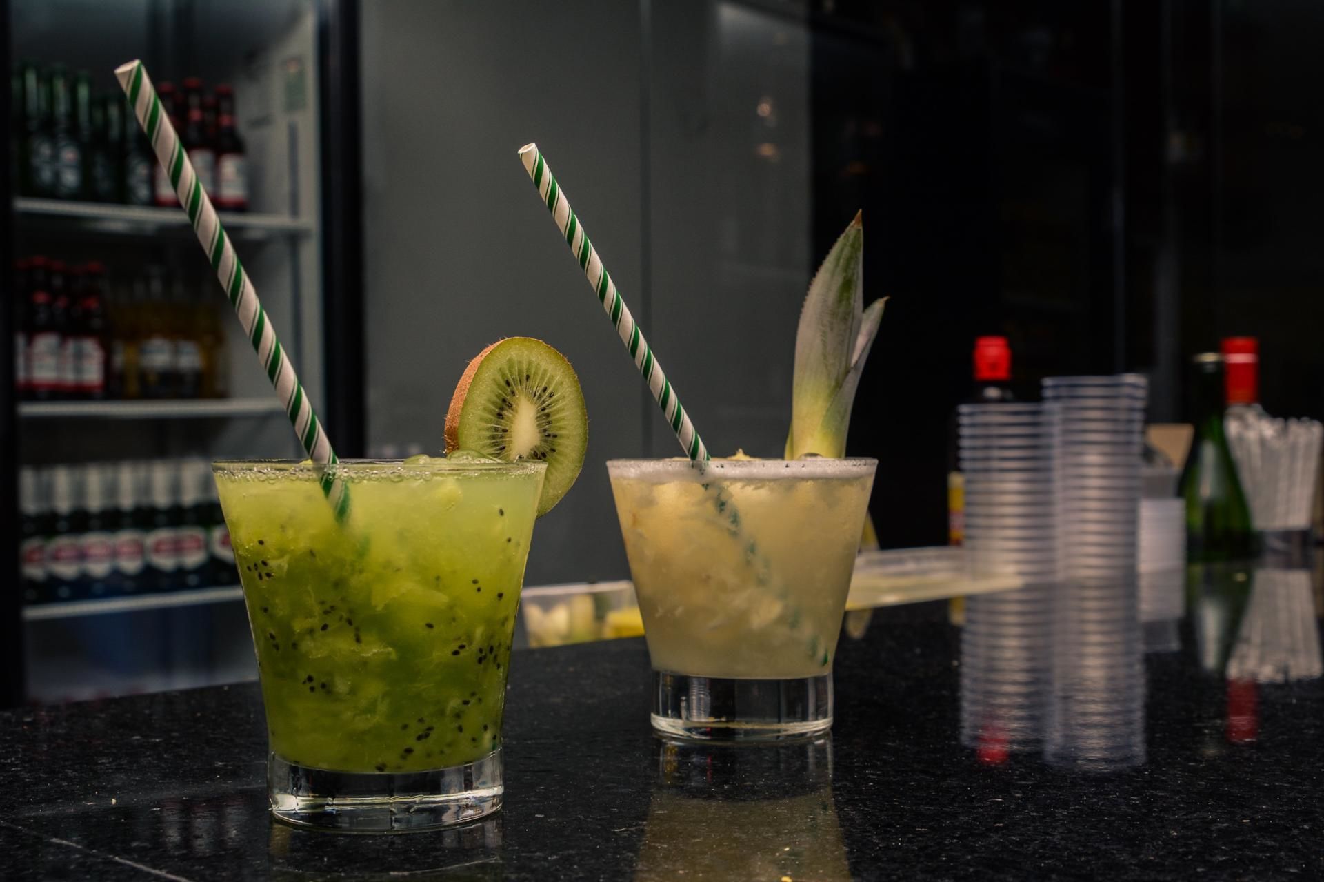 Two cocktails with straws and garnishes on a bar counter.