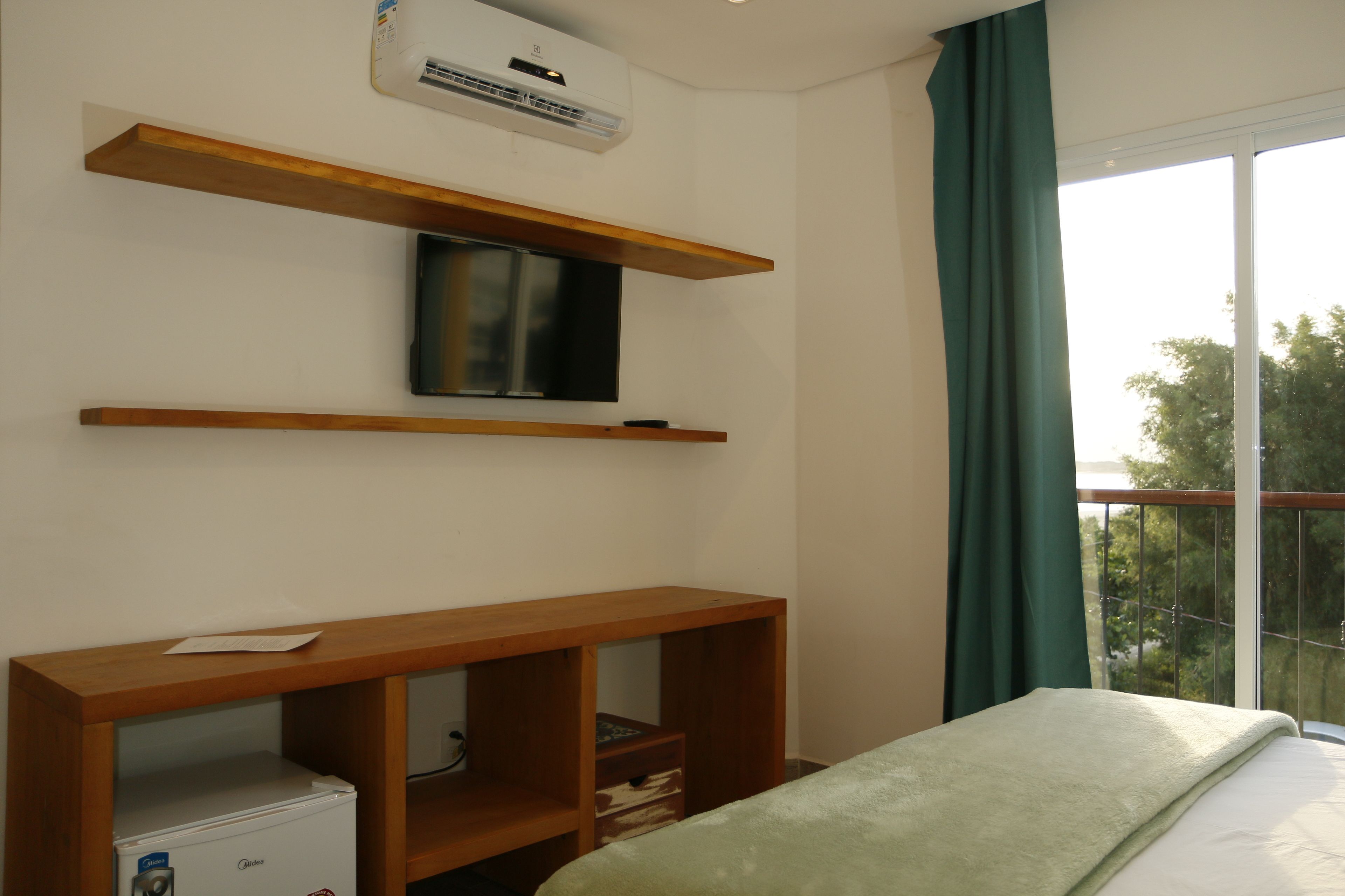 A bedroom with a bed, a TV mounted on a wooden shelf, a small white refrigerator, and a balcony with a view of trees.