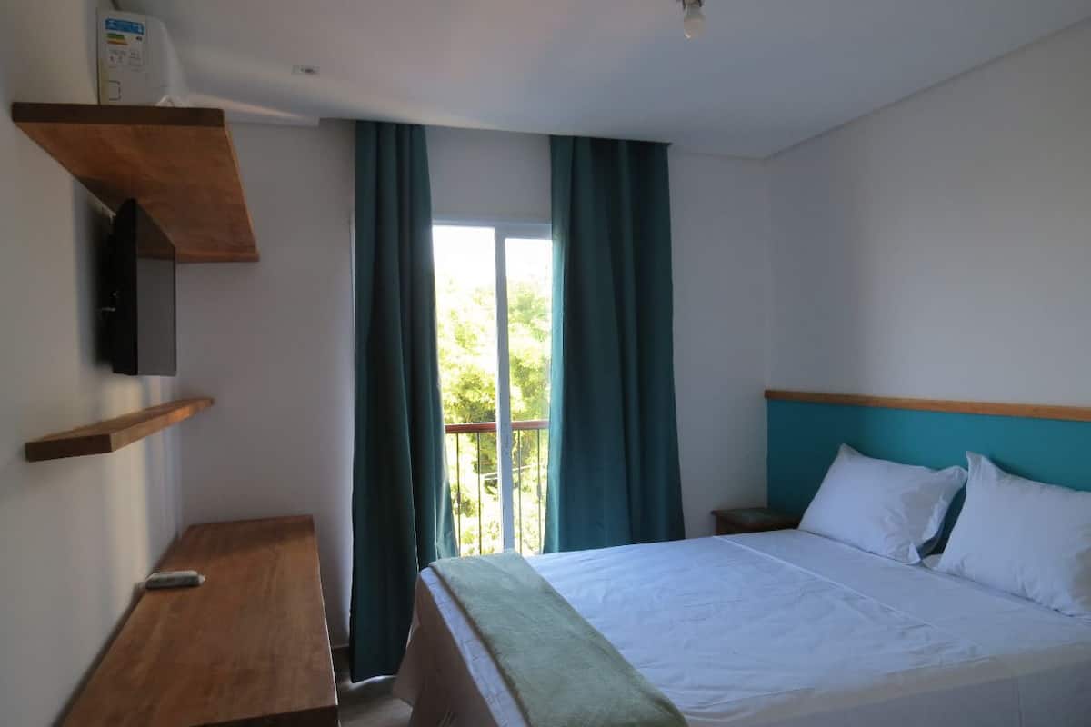 A hotel room with a bed, a wooden nightstand, a TV mounted on the wall, and a balcony with curtains.