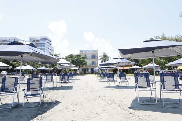 Zona de playa con filas de sillas y sombrillas, un edificio con un cartel que dice "POUSADA", y un cielo despejado.