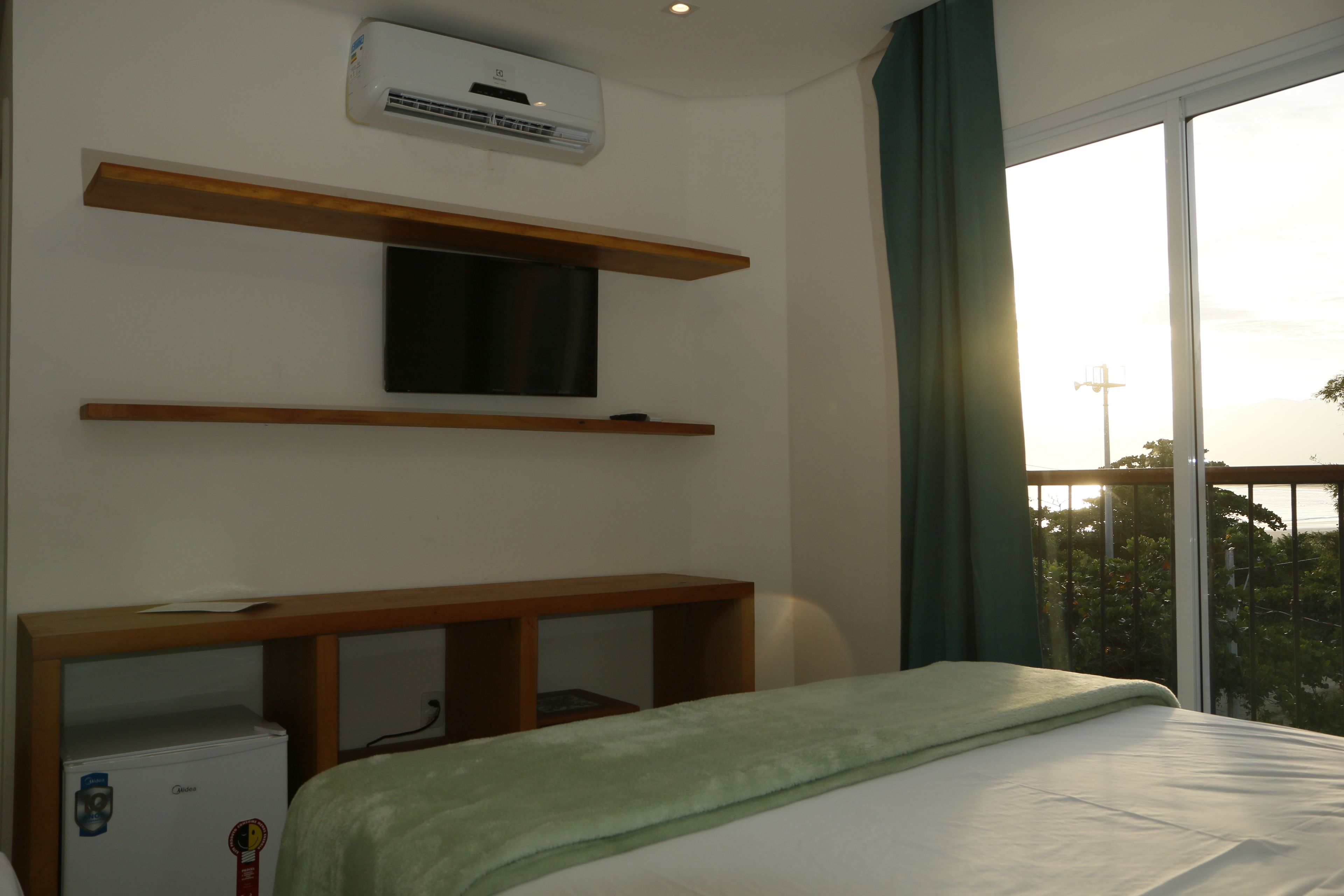 Una habitación de hotel con una cama, un televisor montado en un estante de madera, un refrigerador pequeño y una ventana con vista a follaje.