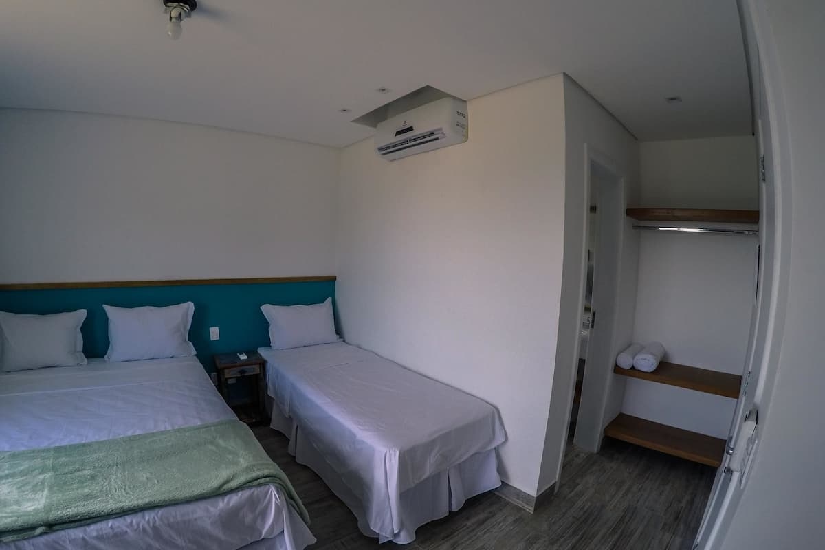 A hotel room with two beds, a ceiling fan, an air conditioning unit, a wooden shelf with toiletries, and a door leading to another room.