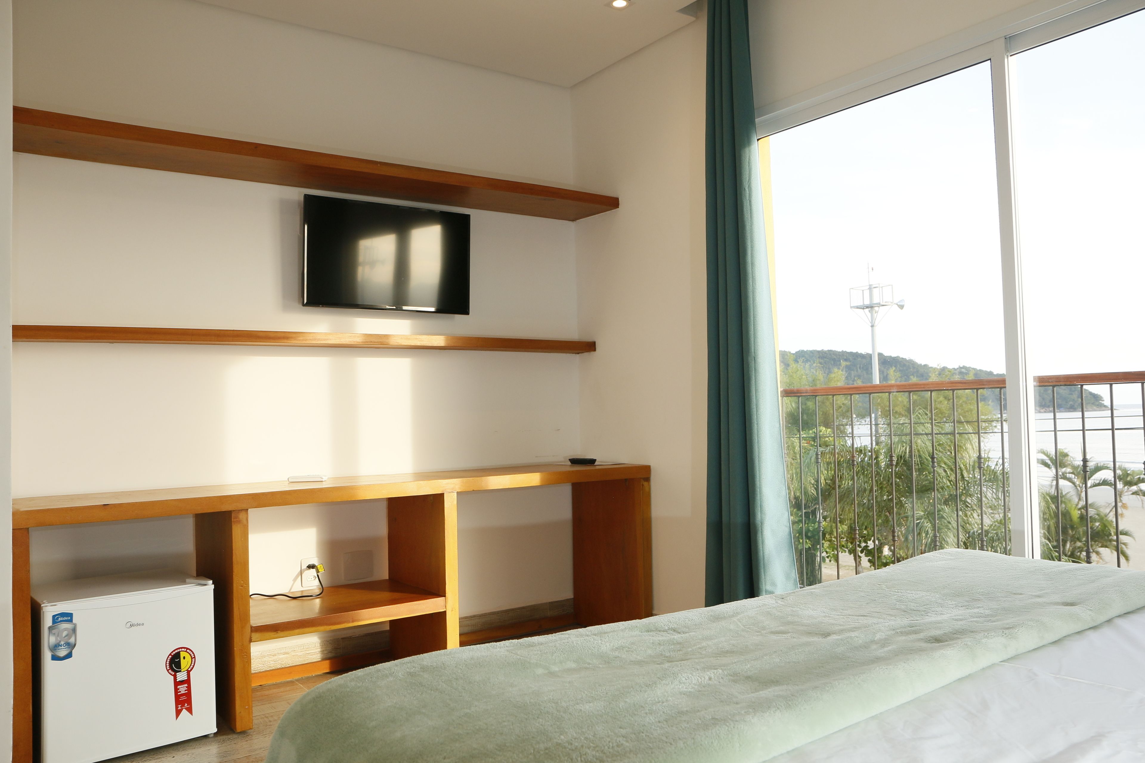 A bedroom with a bed, a TV mounted on a wooden shelf, a small refrigerator, and a window with a view of greenery and hills.