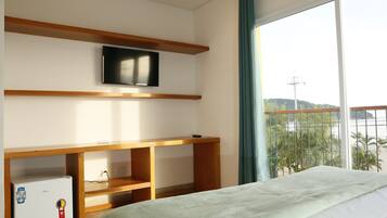 A bedroom with a bed, a TV mounted on a wooden shelf, a small refrigerator, and a window with a view of greenery and hills.