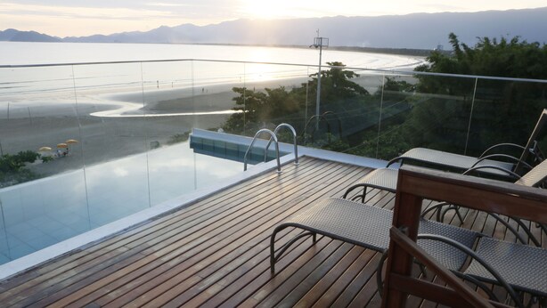 An infinity pool with a wooden deck and metal chairs, overlooking a beach and mountains. - Pousada Conca di Mare (Bertioga)