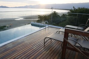 Una piscina infinita con terraza de madera y sillas de metal, con vista a una playa y montañas.