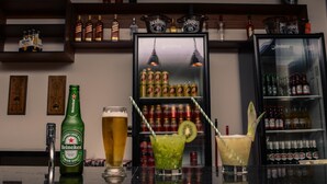 A bar area with a Heineken bottle, a tall beer glass, and two cocktails with straws. Shelves behind display various bottles and cans. - Pousada Conca di Mare (Bertioga)
