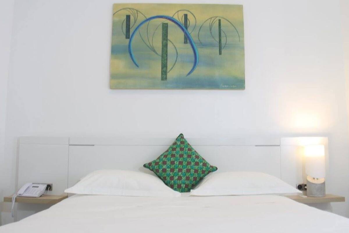 A neatly made bed with white linens, a green patterned pillow, a bedside phone, and a wall-mounted artwork above.