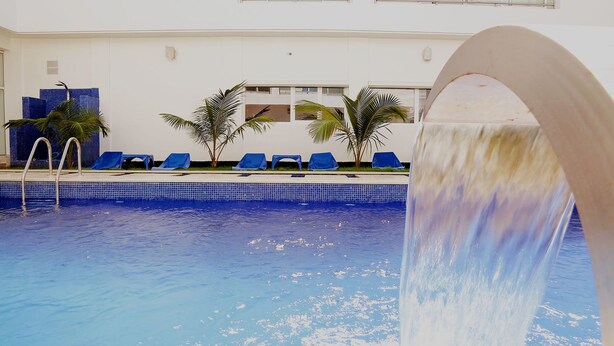 Una piscina con fuente, sillas de descanso azules y palmeras.