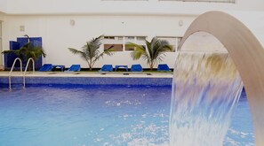 A swimming pool with a water feature, blue lounge chairs, and palm trees.