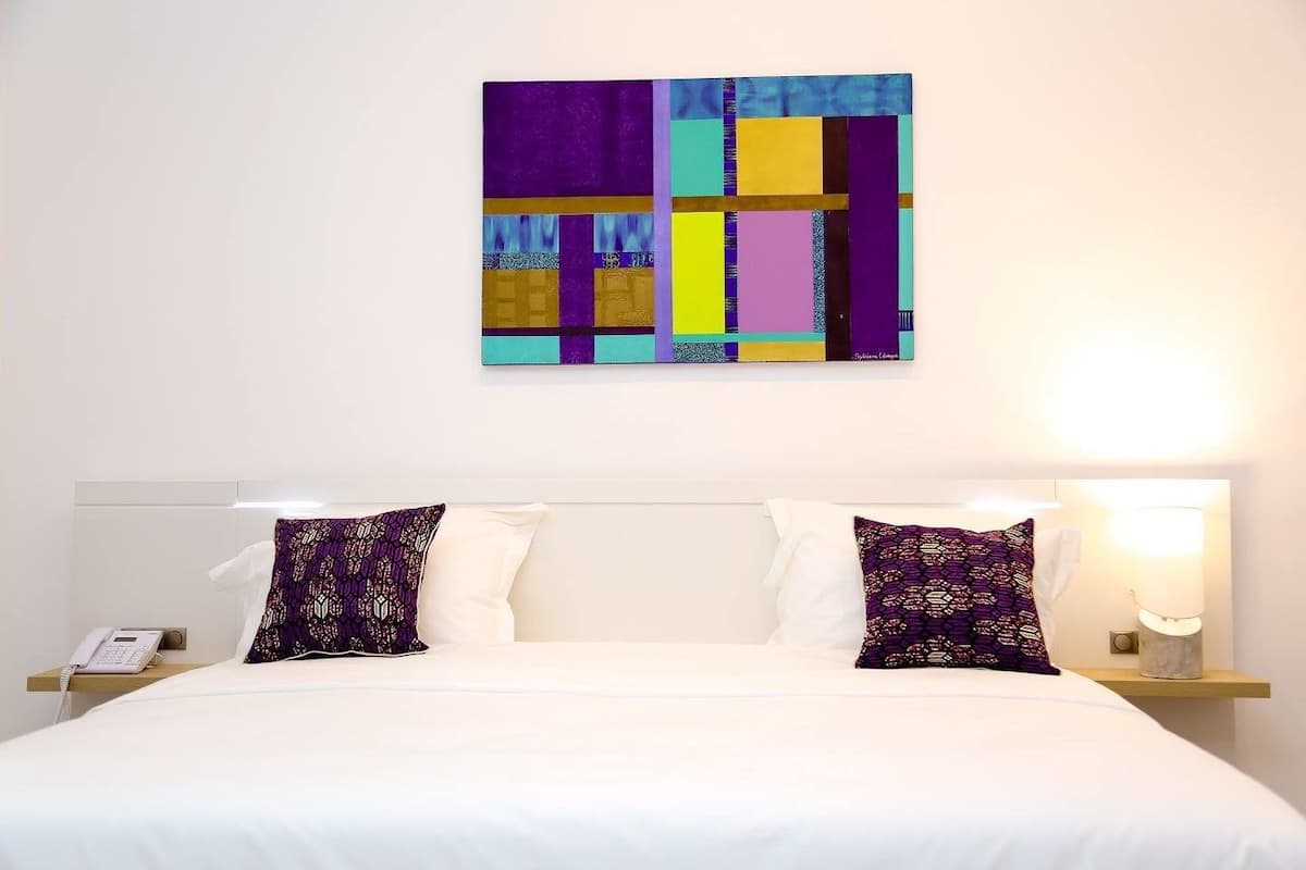 A hotel room with a large bed, two pillows, a bedside table with a lamp, and a wall-mounted artwork.