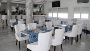 Un restaurante con sillas y mesas blancas, una zona de bar al fondo y una araña.