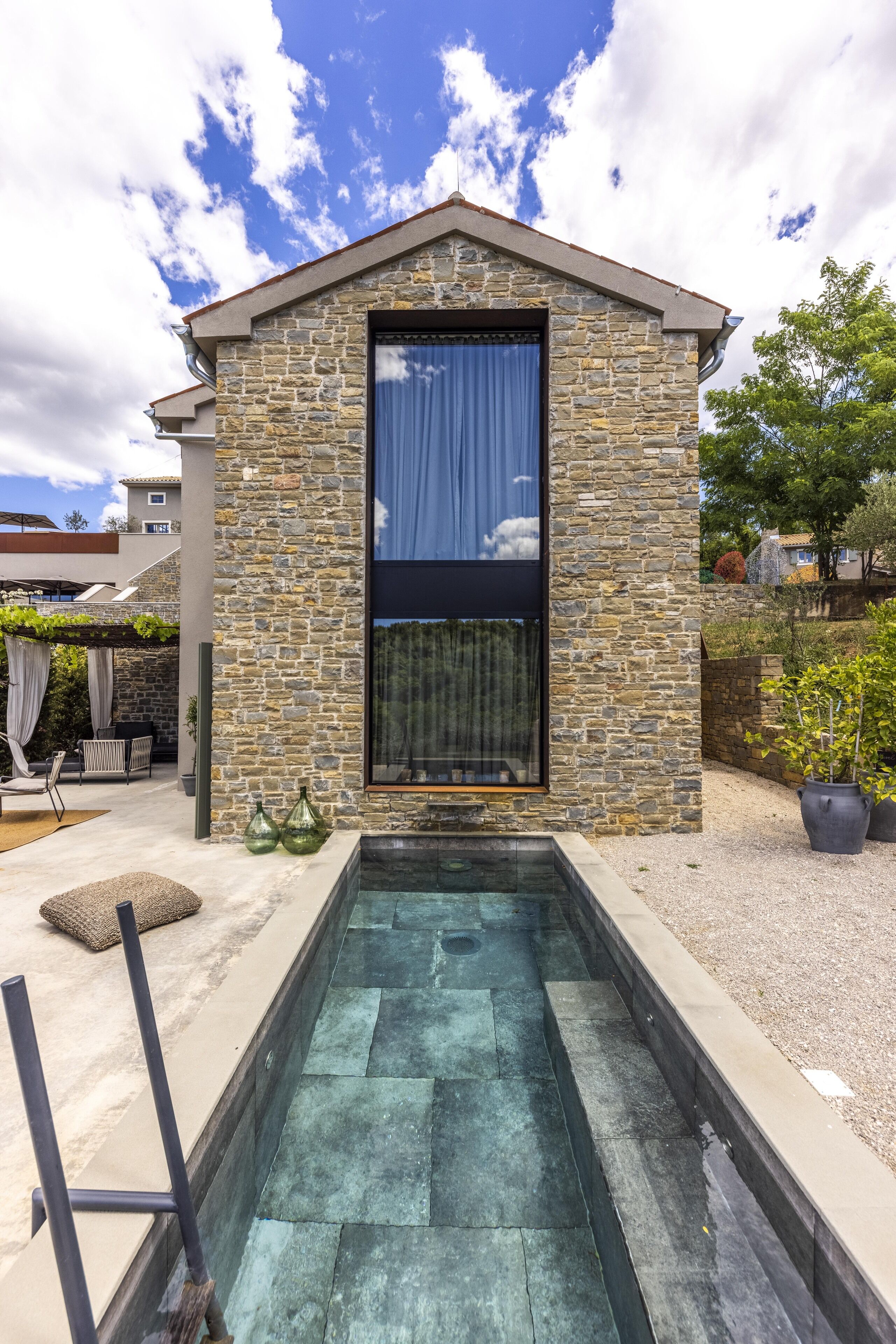 A stone building with a large window, a pool, and outdoor furniture.