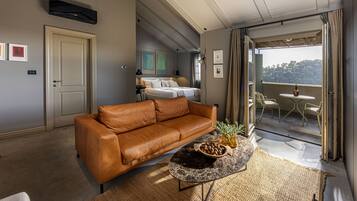 A modern living room with a leather sofa, a coffee table with a bowl of nuts, a bed with white linens, and a balcony with a view of the forest.