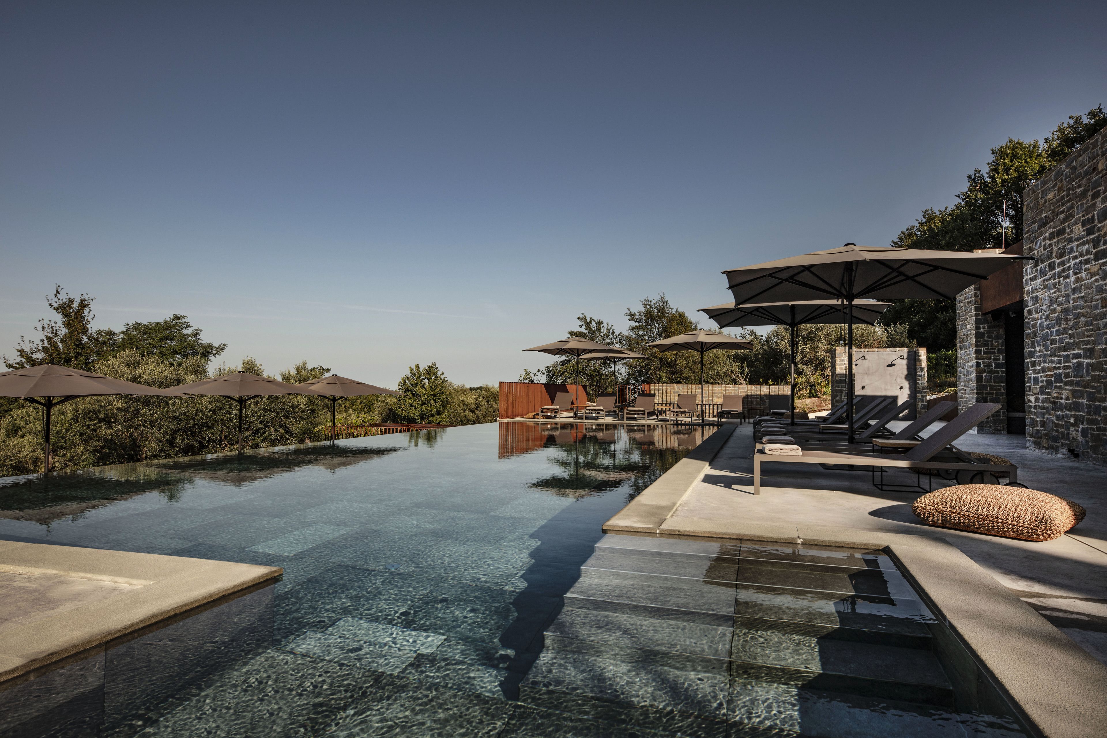 An infinity pool with lounge chairs and umbrellas, surrounded by stone walls and greenery.