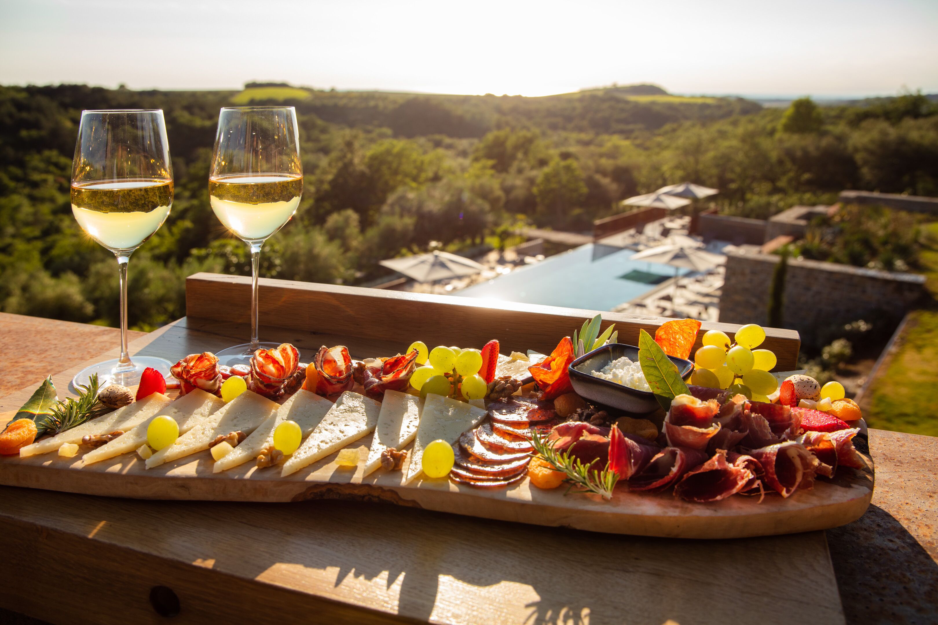 A charcuterie board with various meats, cheeses, and fruits on a wooden table overlooking a pool and lush greenery.