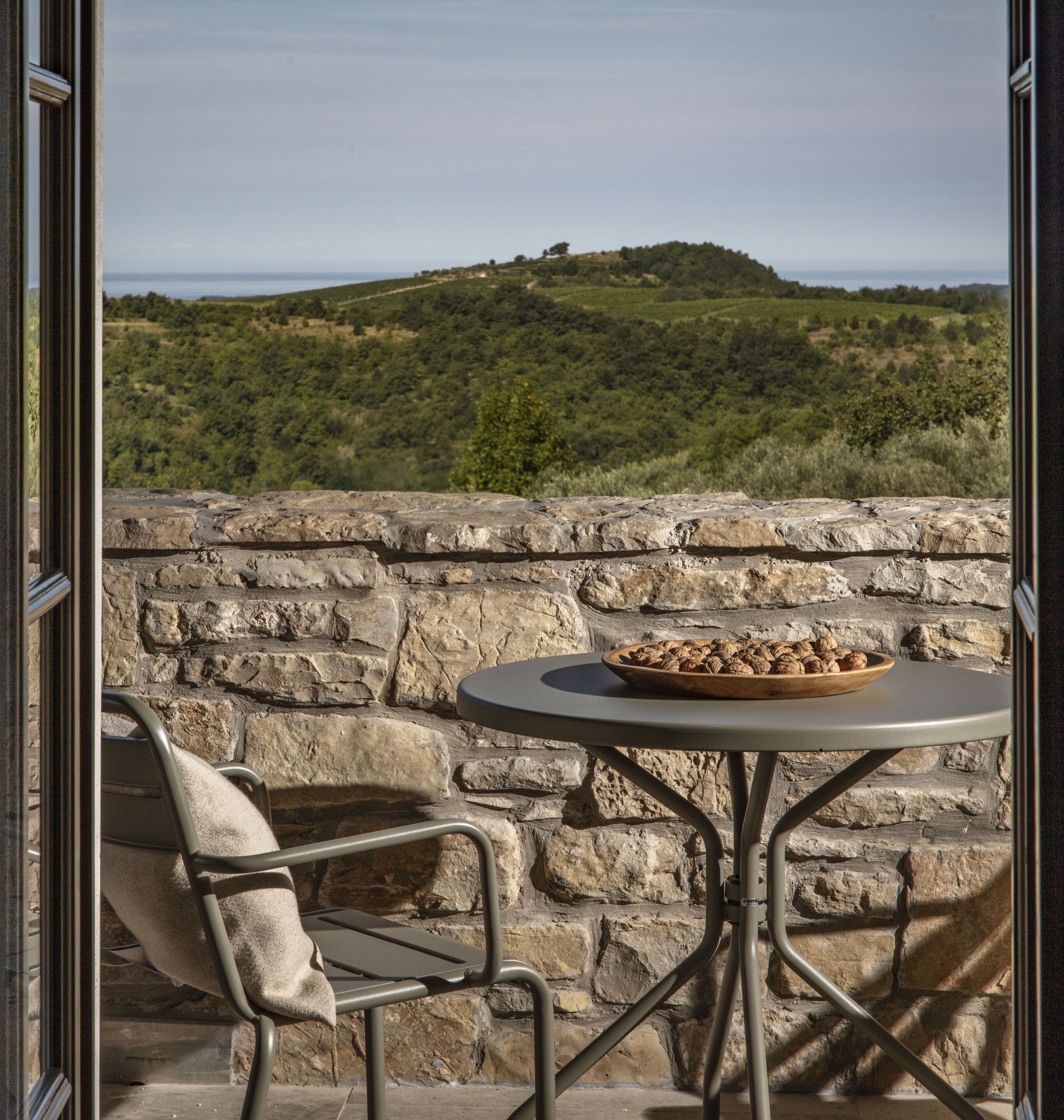 Un muro de piedra con una mesa redonda y una silla, un cuenco de nueces y vistas a una colina verde.