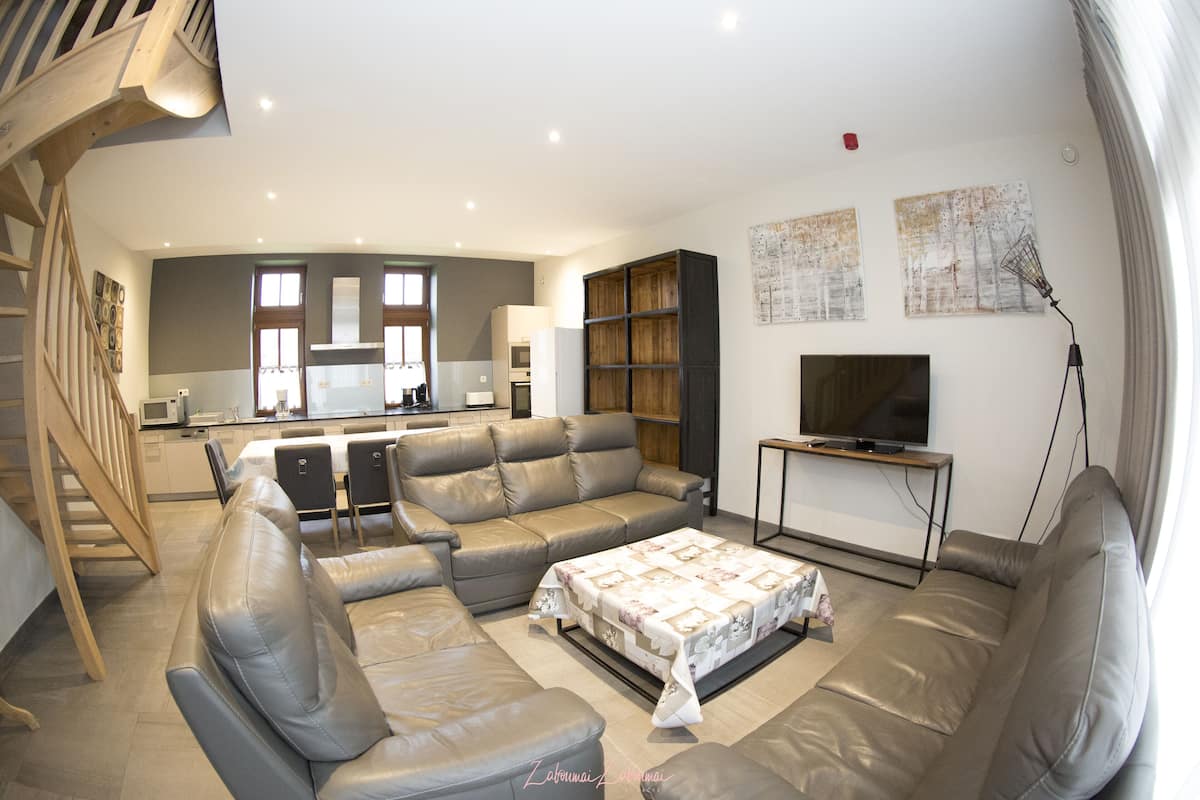 Ein modernes Wohnzimmer mit einer Couch, einem Couchtisch und einem Fernseher. Im Hintergrund ist eine Küche mit einer Mikrowelle und einem Kühlschrank zu sehen.
