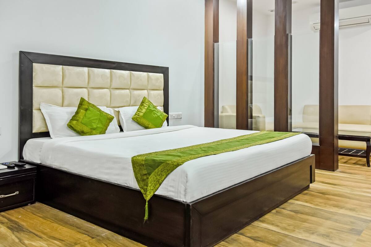 A neatly made bed with a white comforter and green pillows, a wooden nightstand with a remote control, and a wooden floor.