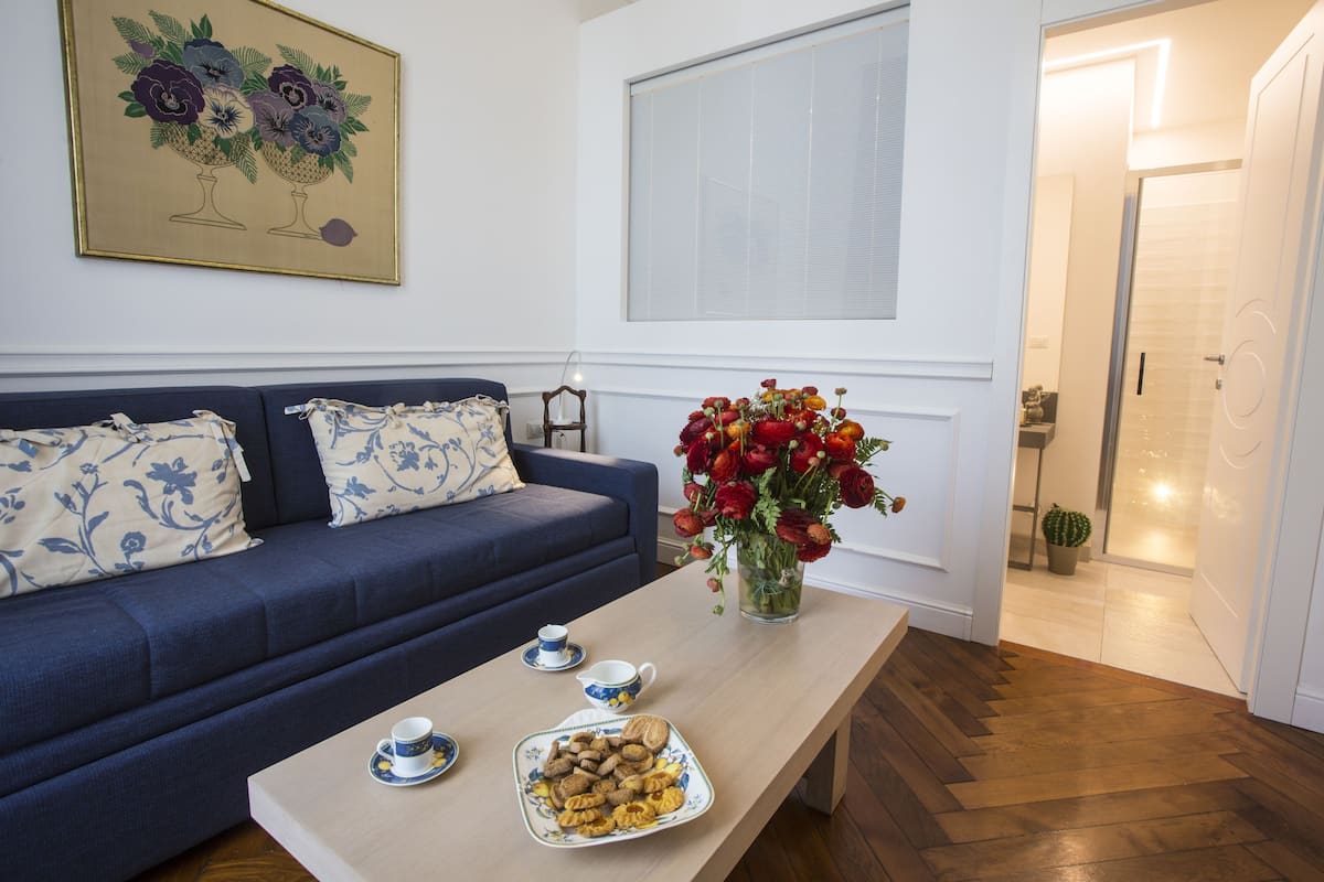 Una sala de estar con un sofá azul, una mesa de centro con un plato de galletas, un florero con flores rojas y un cuadro en la pared.