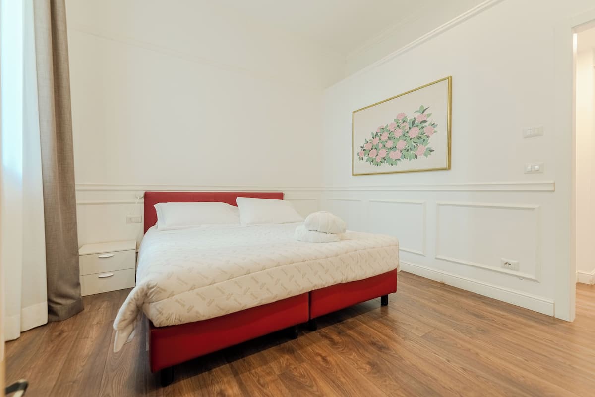 Un dormitorio con una cama roja, paredes blancas, suelo de madera, una mesita de noche blanca y una obra de arte floral enmarco.