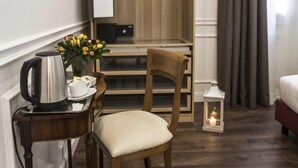 Habitación de hotel compacta con suelo de madera, un escritorio con hervidor, una silla, una mesita con un jarrón de flores, una cama, un armario y una ventana con cortinas.
