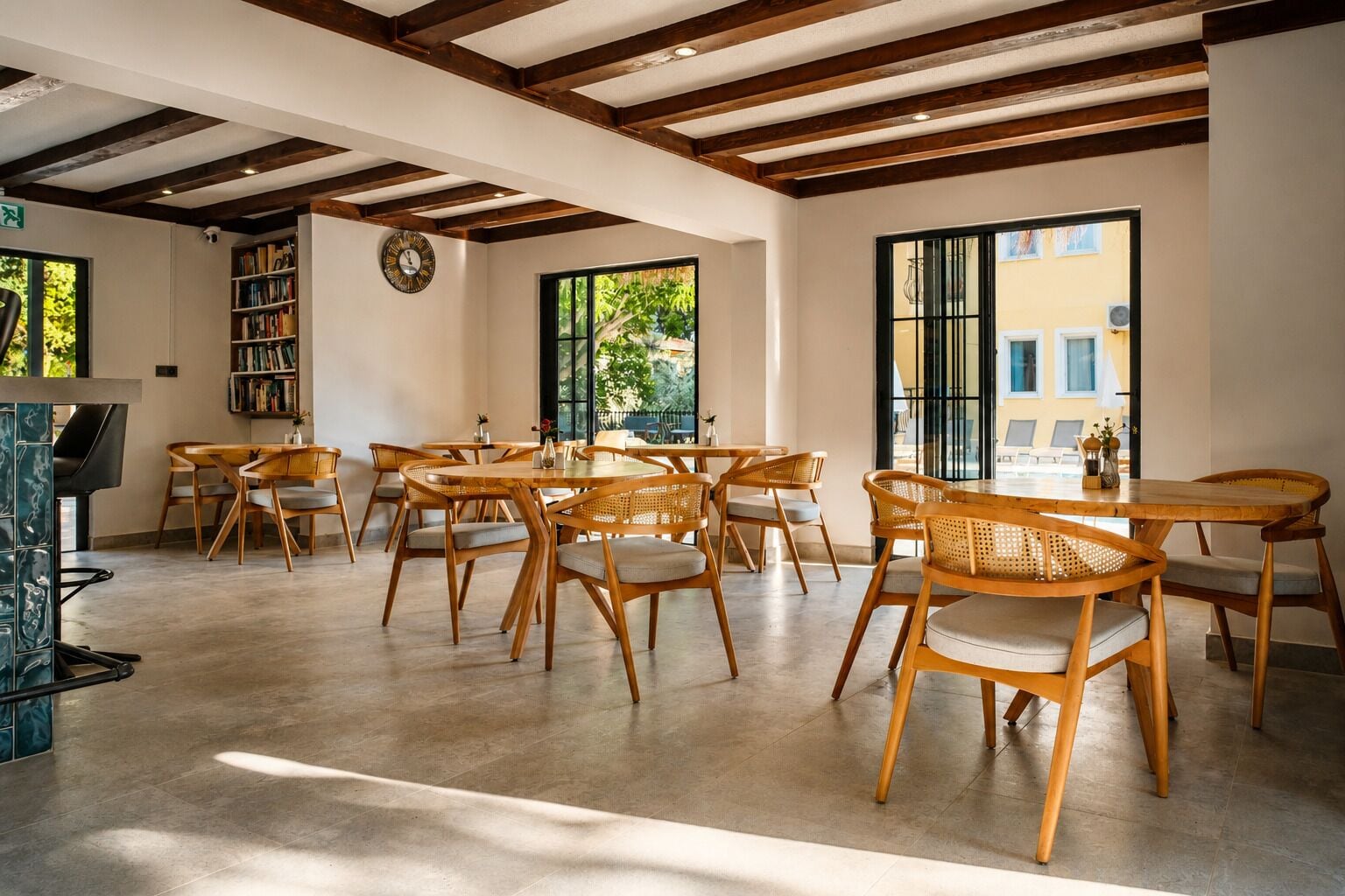 A spacious dining area with wooden tables and chairs, a bookshelf, and large windows with a view of greenery.