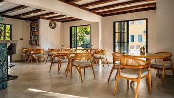 Uma sala de jantar espaçosa com mesas e cadeiras de madeira, uma estante de livros e grandes janelas com vista para o verde.