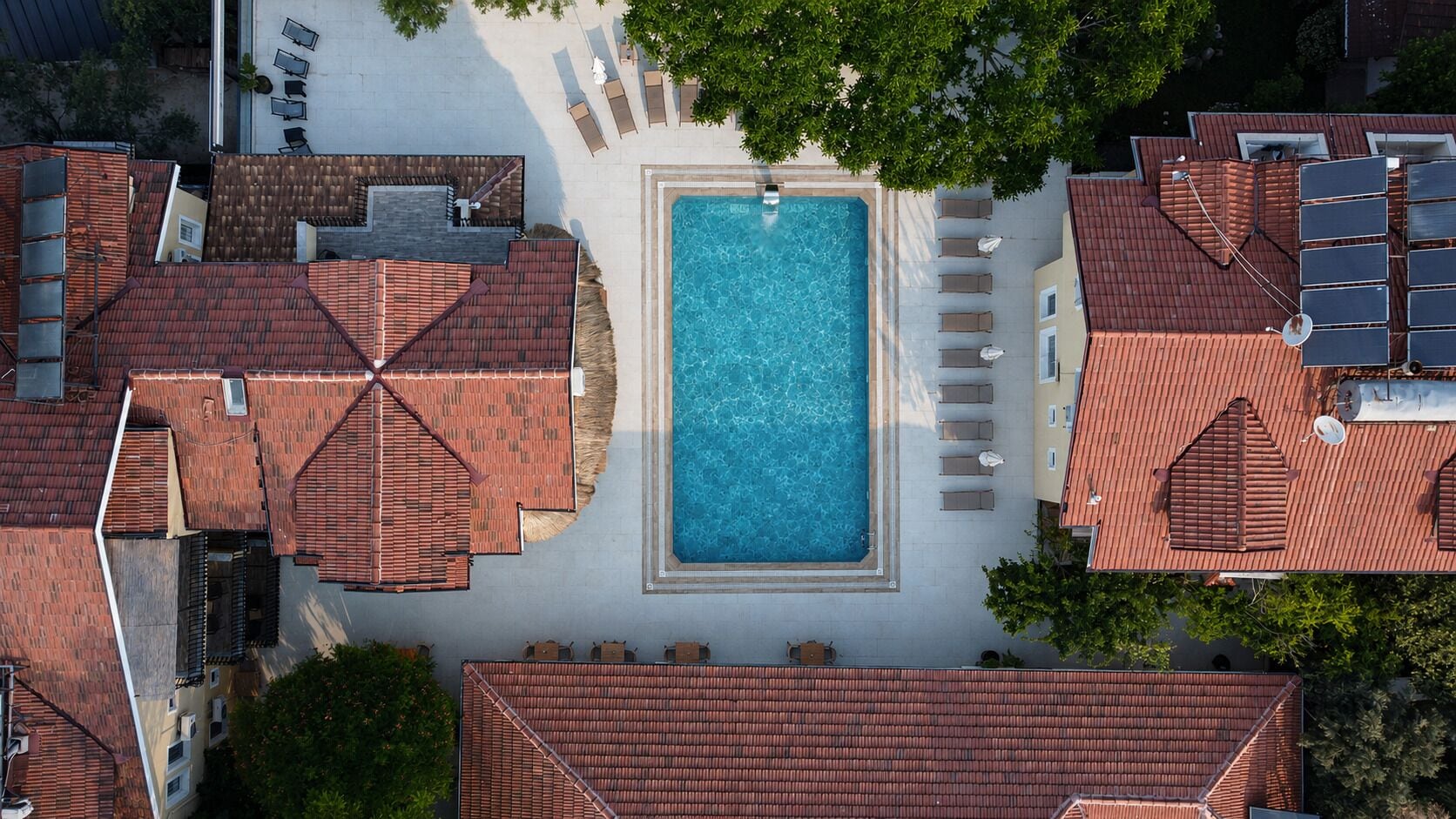 Vue aérienne d’un quartier résidentiel avec une piscine centrale entourée de chaises longues et de parasols, des toits recouverts de tuiles rouges et de la verdure.