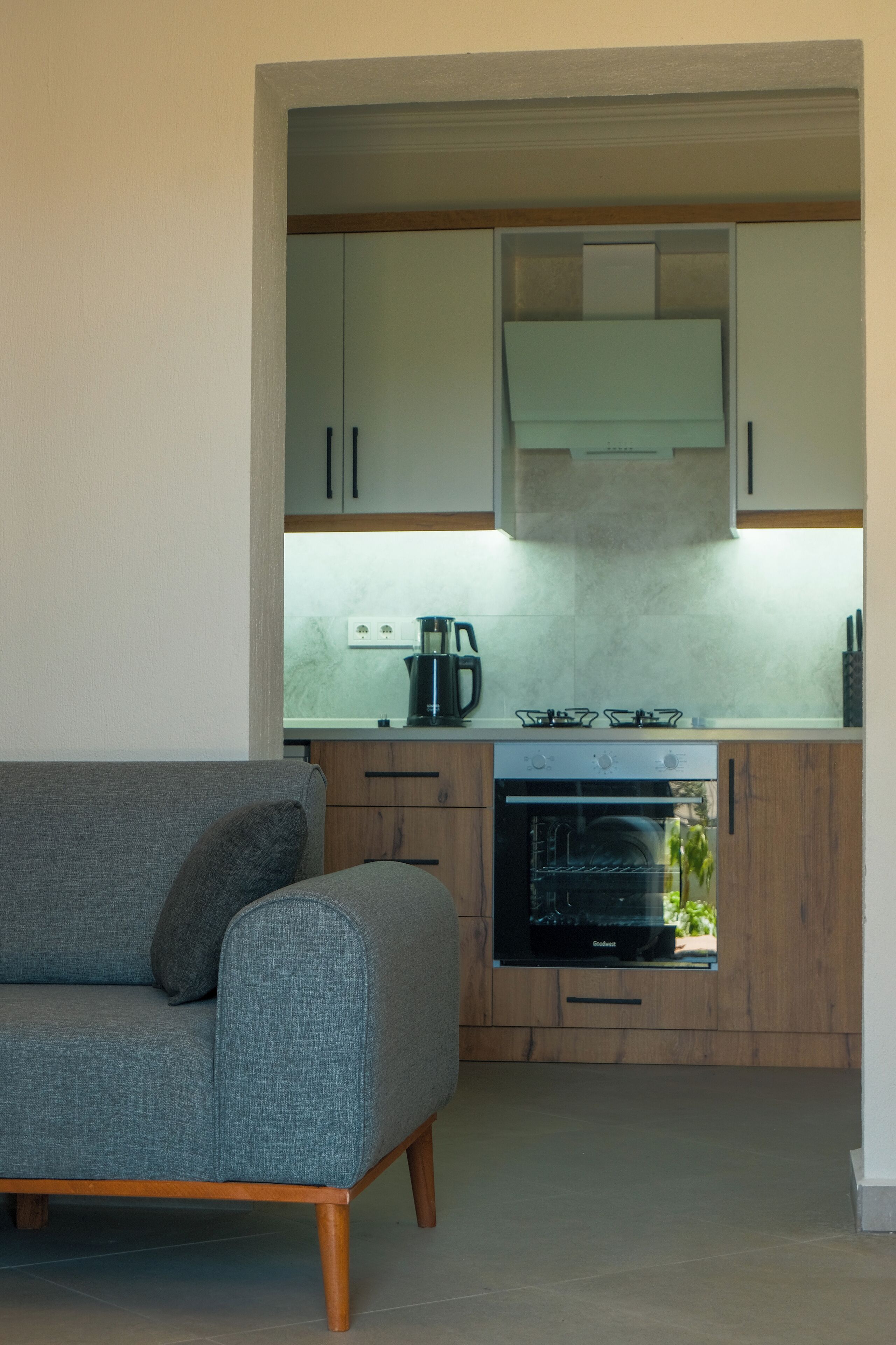 A compact kitchen with wooden cabinets, a built-in oven, and a grey sofa in a corner.