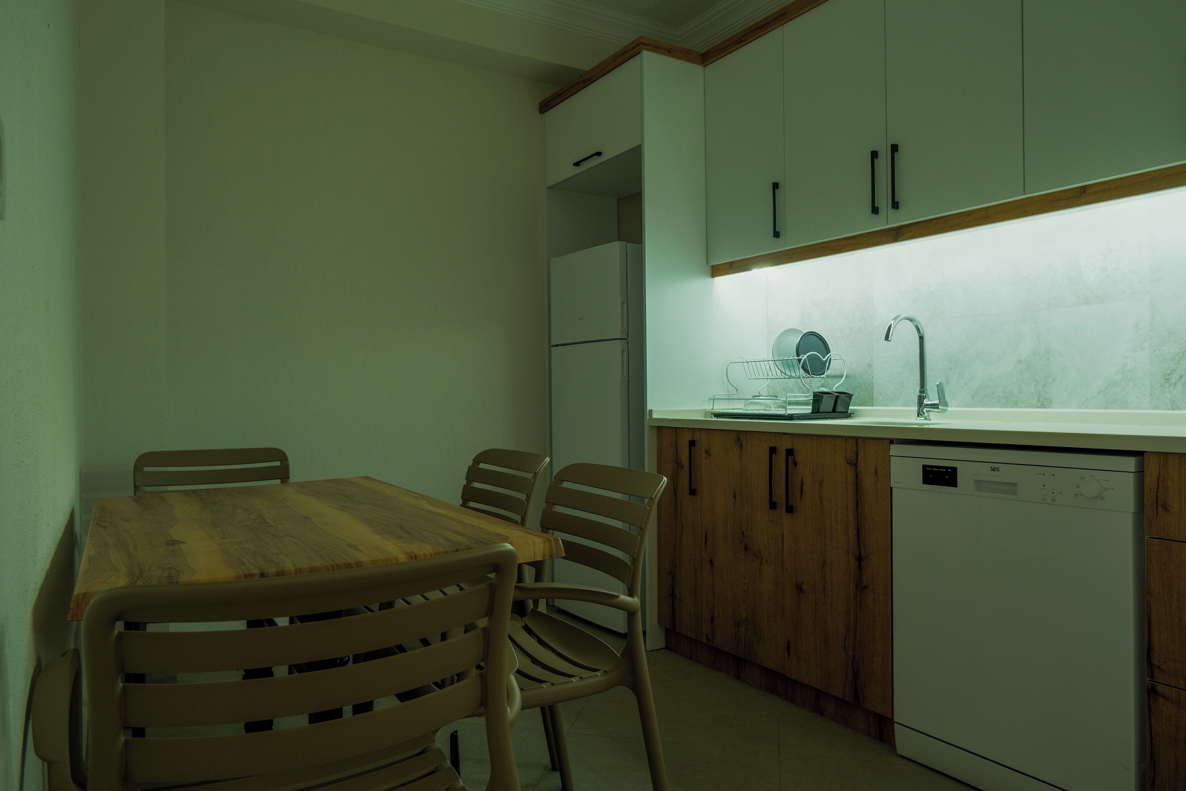Una cocina compacta con una mesa y sillas de madera, armarios blancos, lavavajillas integrado y salpicadero de mármol.