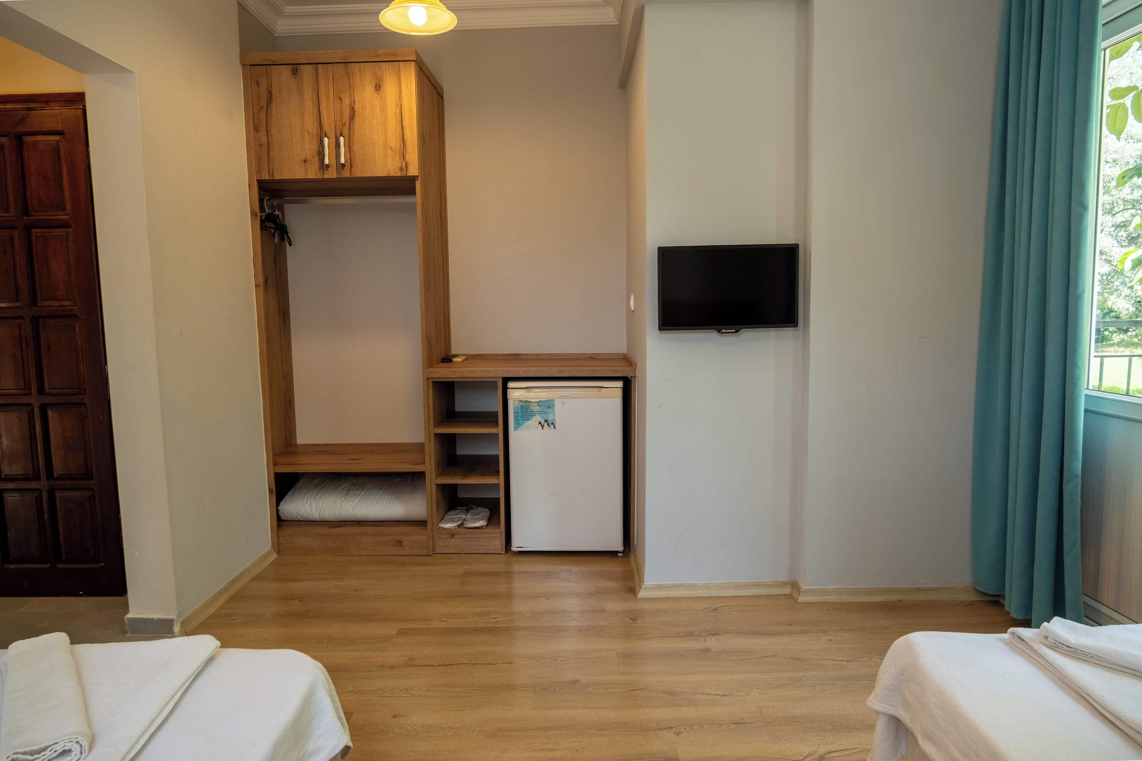 Quarto pequeno e limpo com piso de madeira, uma cama branca, um refrigerador branco compacto, um guarda-roupa de madeira e uma TV fixada na parede.
