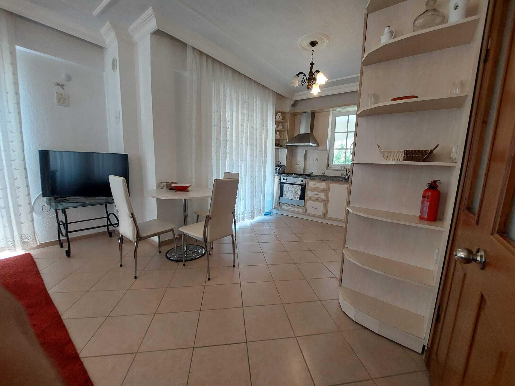 A dining area with a TV, chairs, and a table. There is a kitchen area with cabinets and a stove.