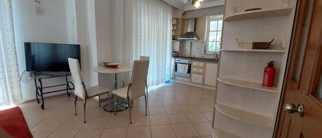A dining area with a TV, chairs, and a table. There is a kitchen area with cabinets and a stove.